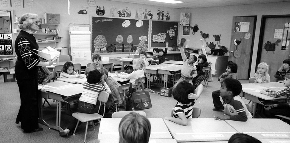 Black and white photograph of a classroom at Glen Forest Elementary School in November 1981. A teacher is leading a class of what look to be second or third graders. The children are very engaged in the lesson and have their hands up in the air waiting to answer.